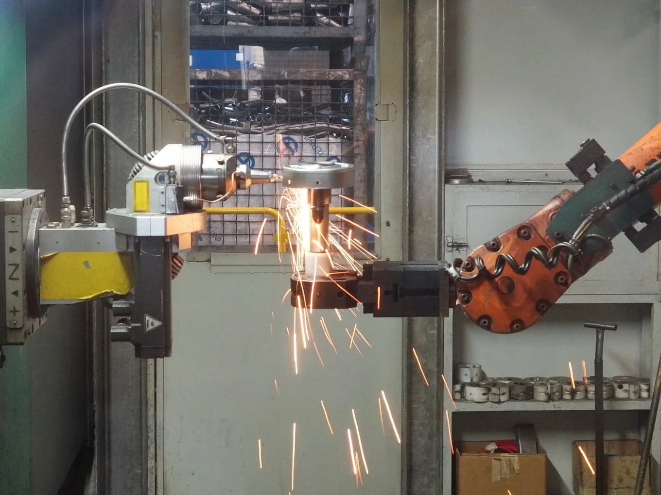 Industrial robot arm holding a metal part at a welding station, sparks flying from the weld